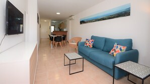 A living room with a blue sofa, two patterned coffee tables, a flat-screen TV mounted on the wall, and a painting of a landscape. - Sa Gavina C, 2 Bedroom apartment with WIFI, POOL, LIFT, balcony or terace. (L'Estartit)