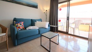 A living room with a blue sofa, a small table, and a view of the outside balcony. - Sa Gavina C, 2 Bedroom apartment with WIFI, POOL, LIFT, balcony or terace. (L'Estartit)