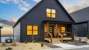 Une maison moderne à deux étages, dotée d’une façade sombre, d’un porche avec des marches en bois et d’une porte bien visible.