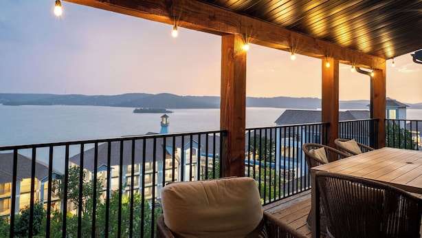 Une terrasse en bois avec une table et des chaises, d’où l’on peut admirer un plan d’eau et des bâtiments.