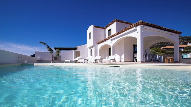 Un espace aménagé au bord d’une piscine à l’eau d’un bleu limpide, avec un bâtiment blanc orné d’arcades et des chaises longues.