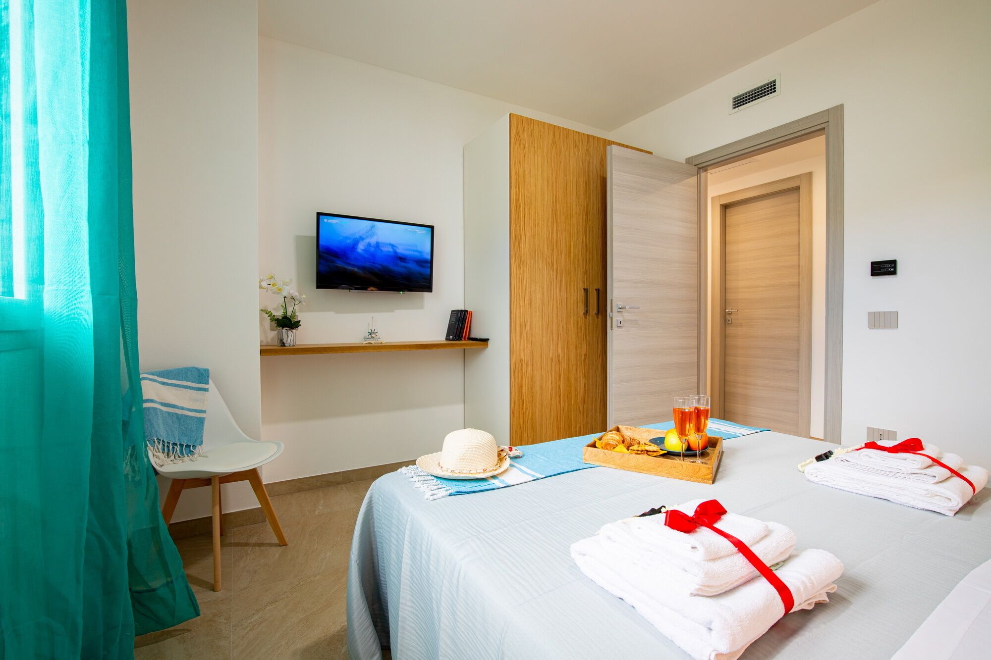 Une chambre d’hôtel comprenant un lit, une télévision, une armoire en bois, une petite table avec des fruits et des verres, et une chaise.