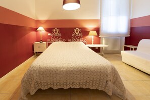 A bedroom with a large bed, two bedside tables with lamps, a white sofa, and a window with sheer curtains. - Family room "The Roses" with private bathroom in the room (Livorno)