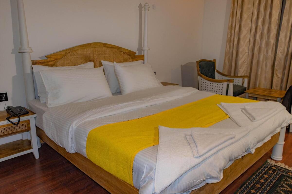 A neatly made bed with a yellow blanket, white pillows, and a wooden headboard. There is a small bedside table with a telephone and a chair with a cushion.