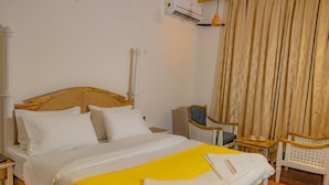 Un lit bien fait, avec une literie blanche et un plaid jaune, une tête de lit en bois, une petite table de chevet avec une lampe et une chaise avec un dossier en osier.