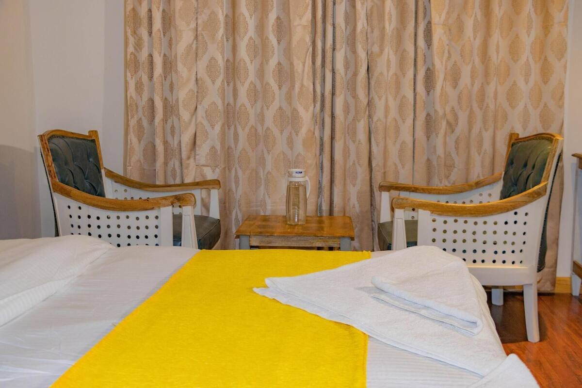 A hotel room with a bed, two armchairs, a small table with a bottle, and patterned curtains.