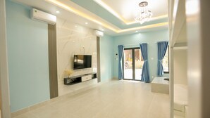 A modern living room with a flat-screen TV, air conditioning units, a chandelier, and a sliding glass door leading to an outdoor area. - Kiwi's Homestay & Cafe (Mui Ne)