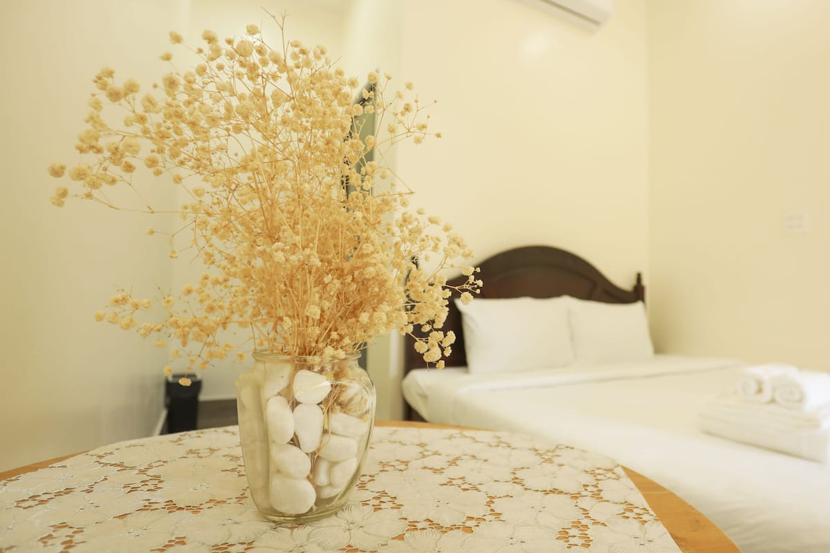 Quarto com cama, mesa de cabeceira e um vaso com flores secas sobre uma mesa.