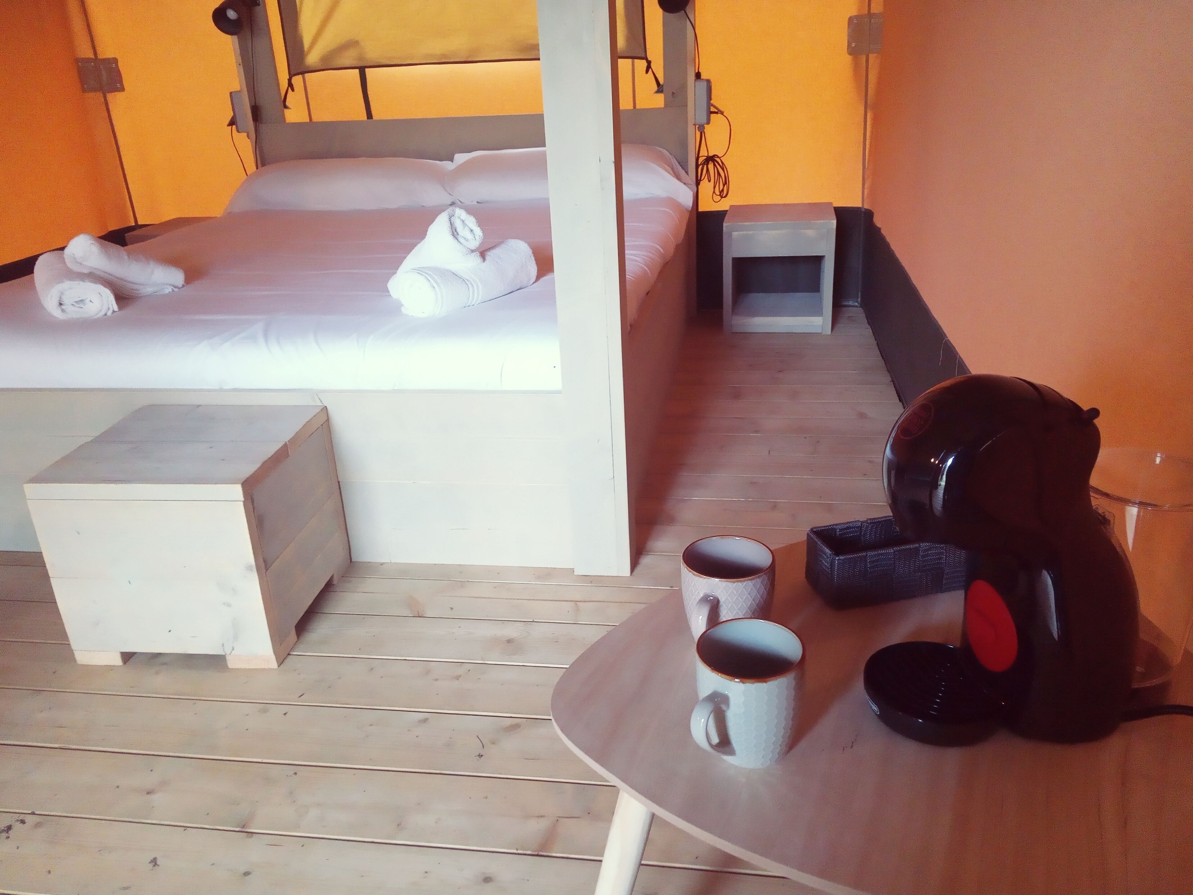 A hotel room with a bed, a bedside table with a lamp, a small table with two mugs, and a pair of headphones on the table.