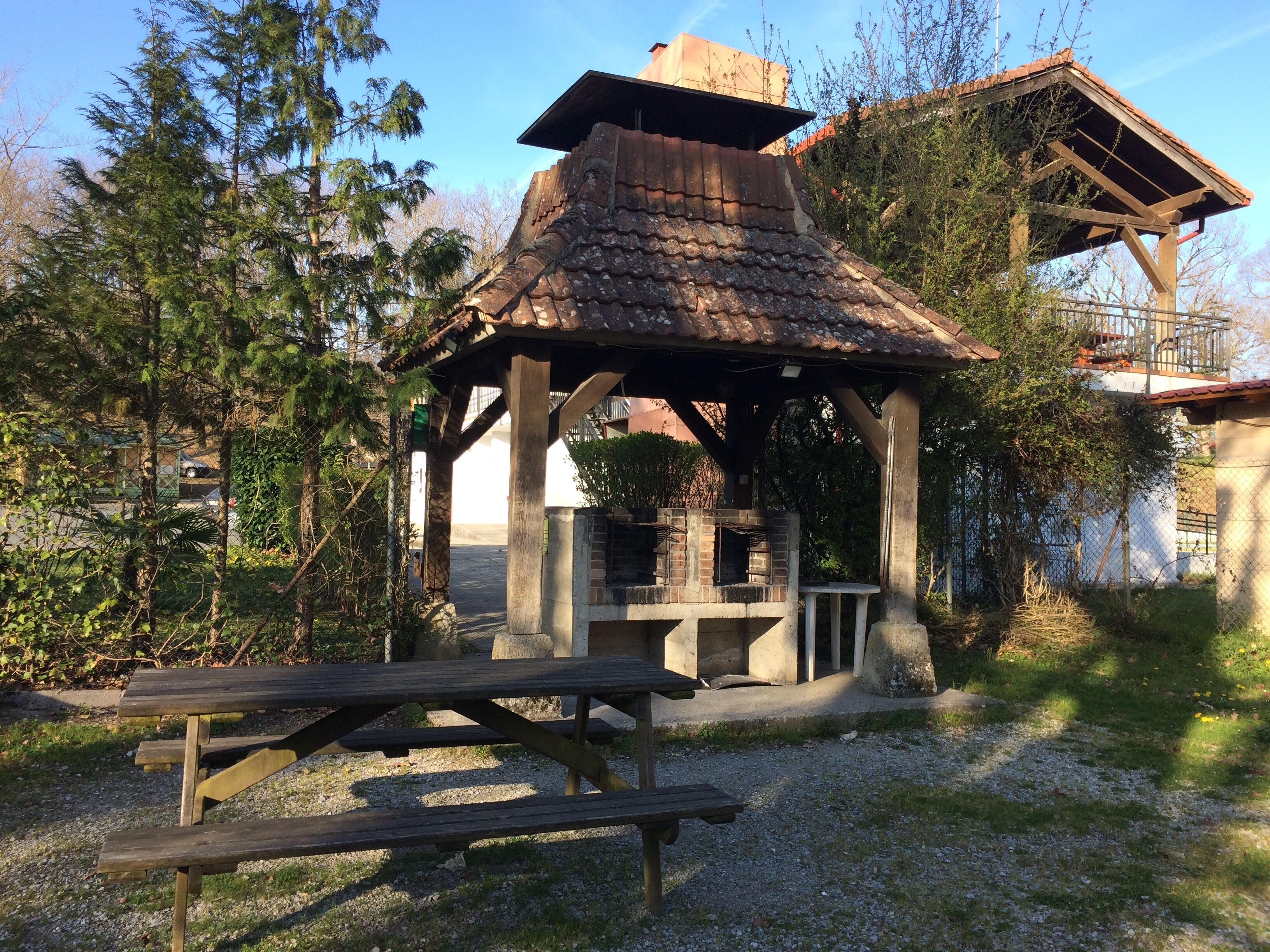 En pavillon med en murstensovn, omgivet af træer og et picnicbord.