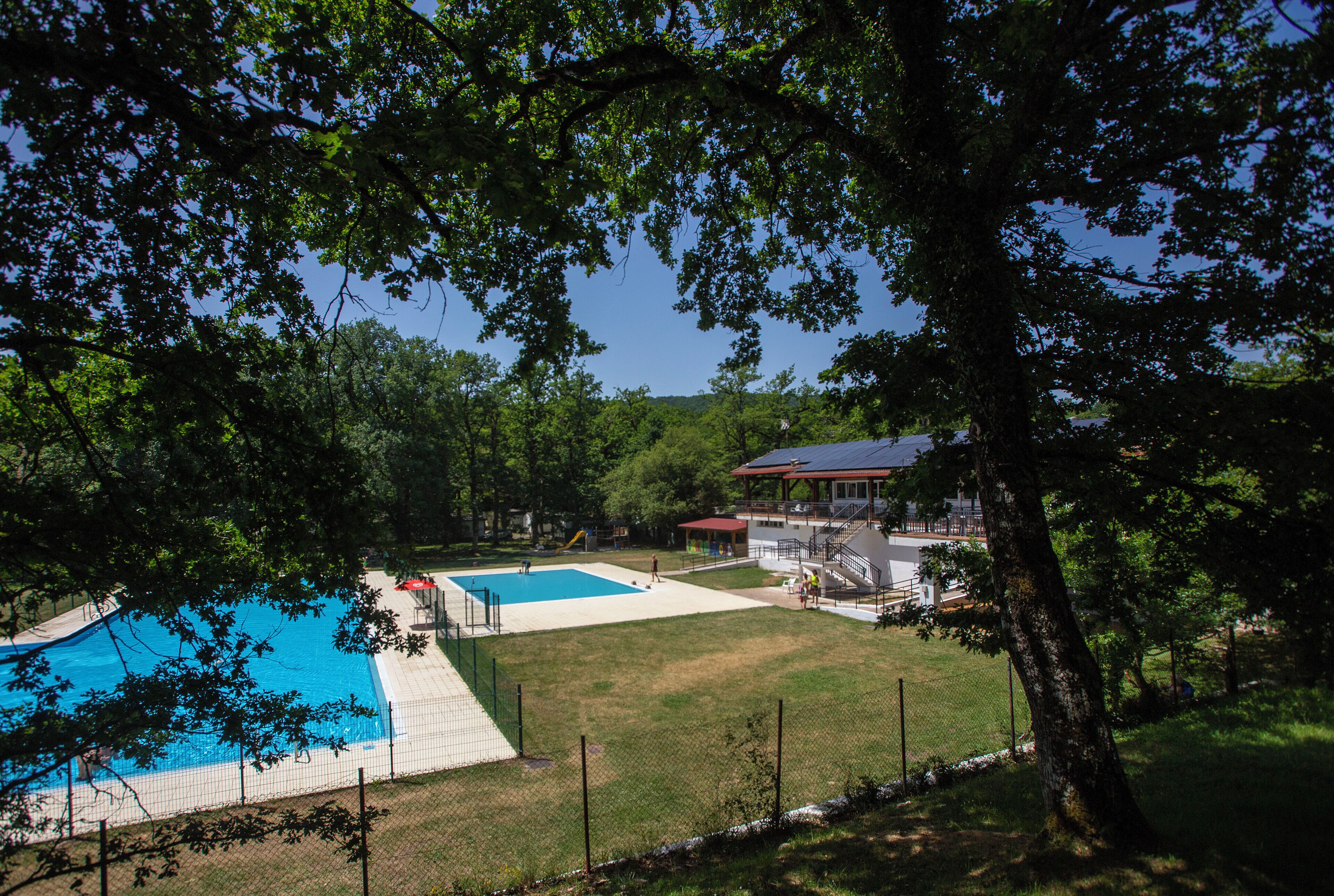 Una piscina con un área de terraza cercada, rodeada de árboles y un edificio con techo rojo.