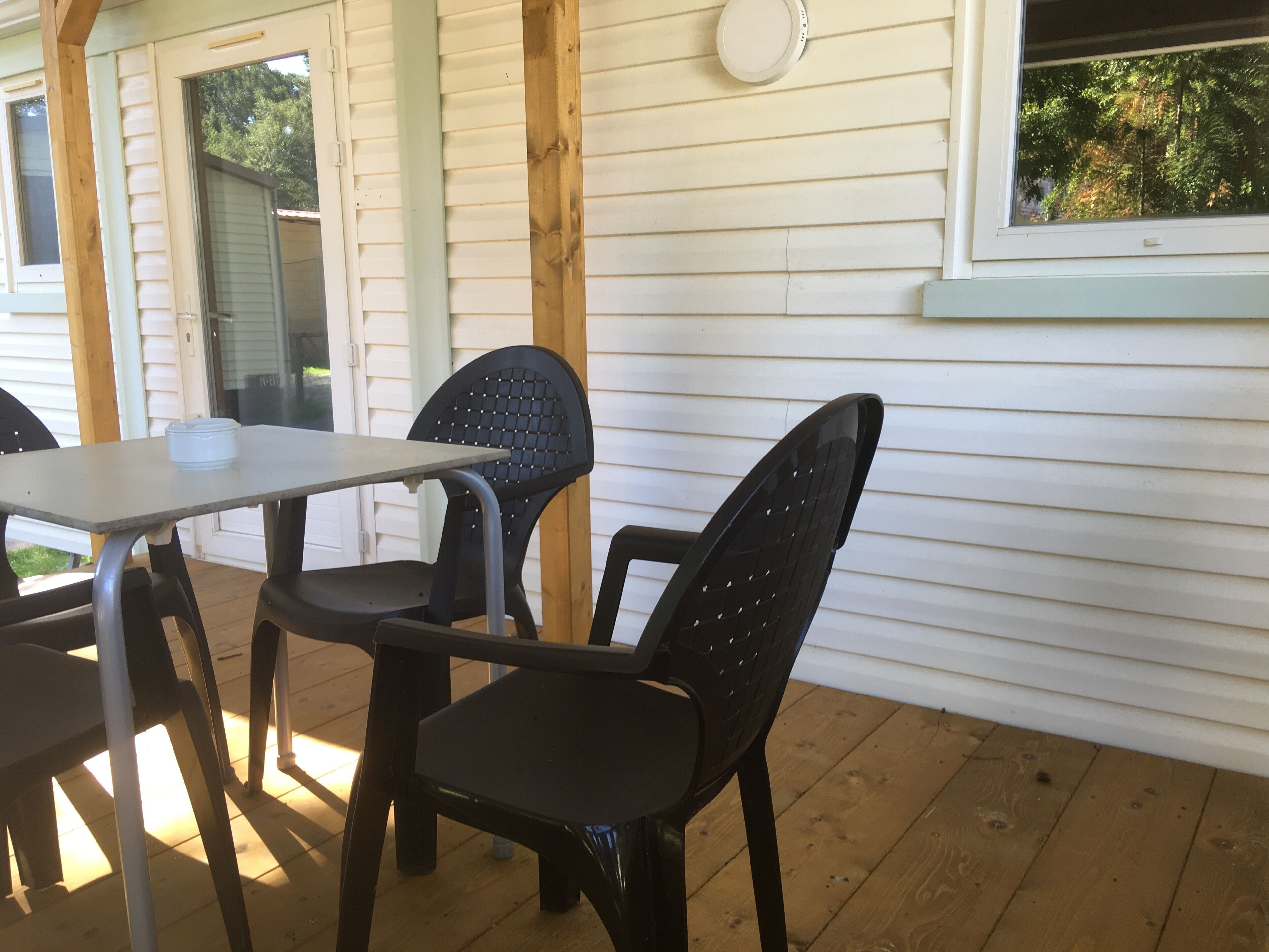A covered patio area with a table and two chairs, wooden deck flooring, and a white exterior wall with horizontal siding.
