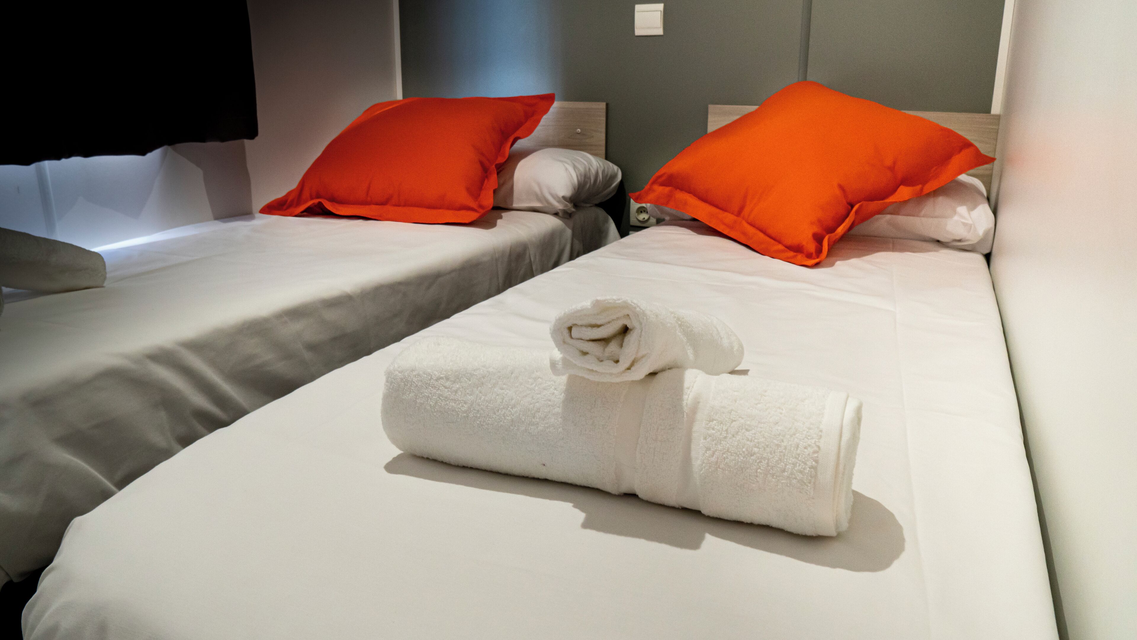 Una camera d'albergo con due letti, ognuno con un materasso bianco e cuscini arancioni. Un asciugamano bianco arrotolato è posizionato sul letto in primo piano.