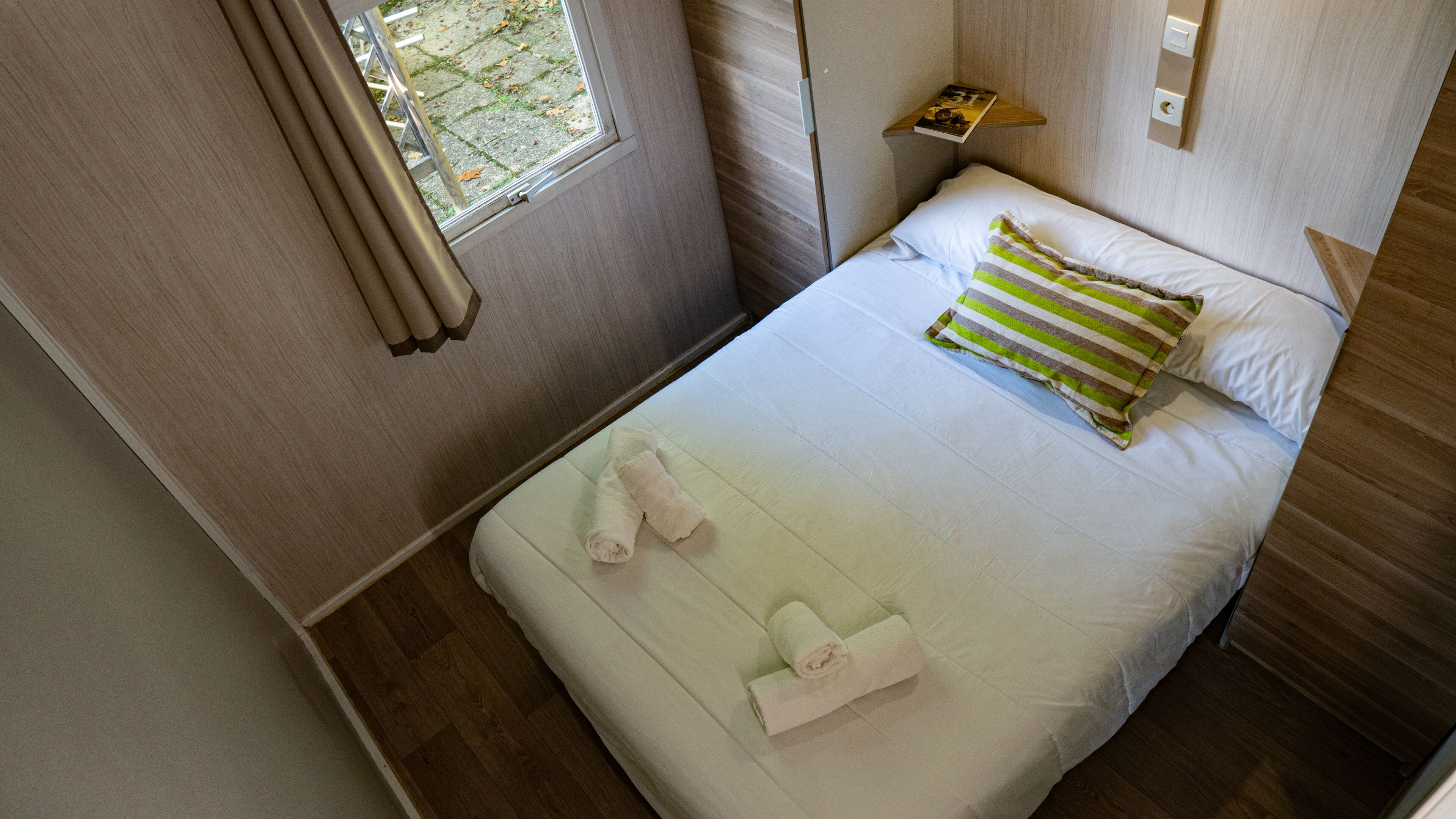 Una cama bien hecha con dos toallas, una ventana con cortinas y una mesita de noche con un libro.