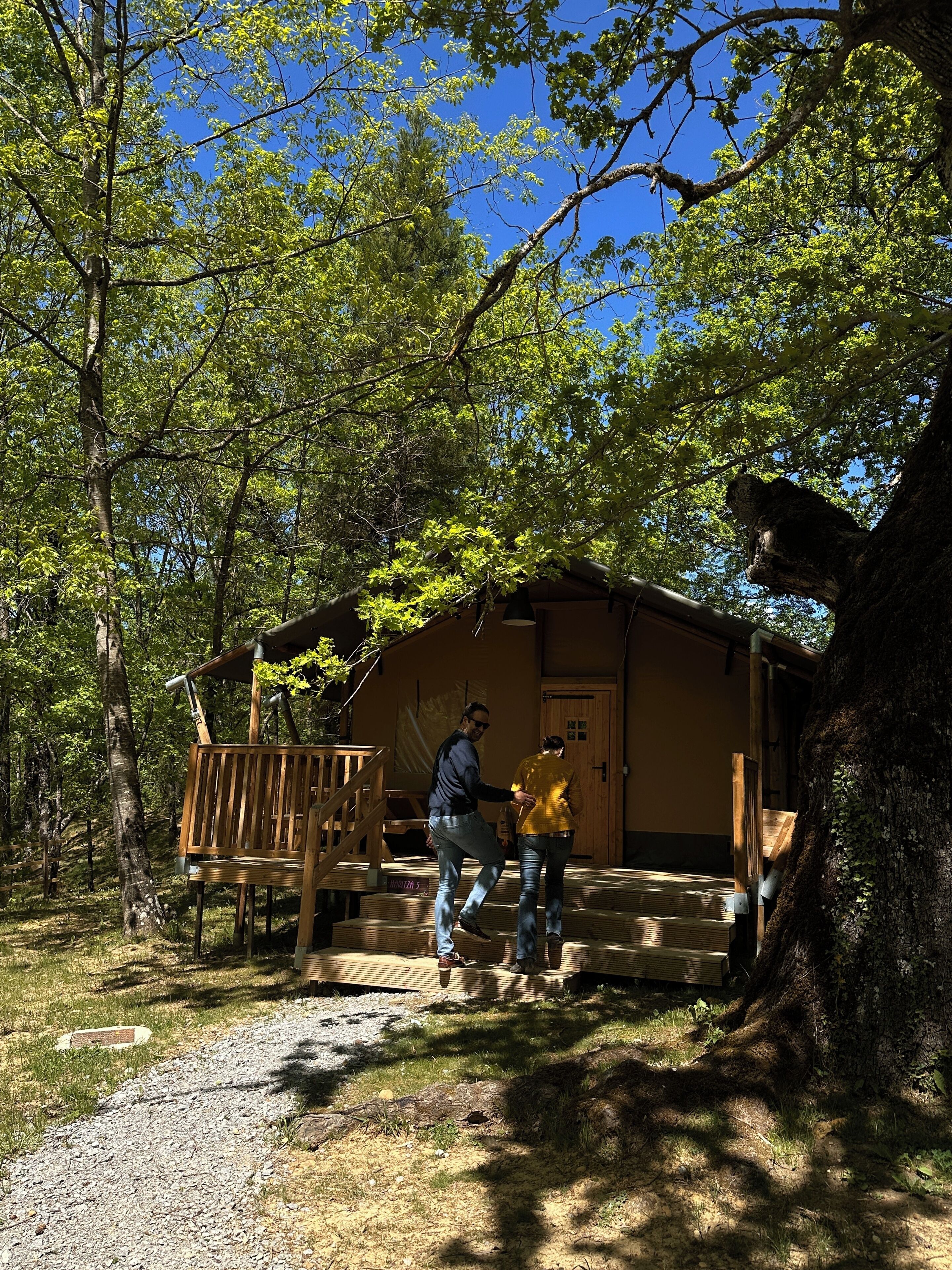 Une cabane dotée d’une terrasse en bois, d’une porte avec une pancarte et d’un grand arbre à proximité.