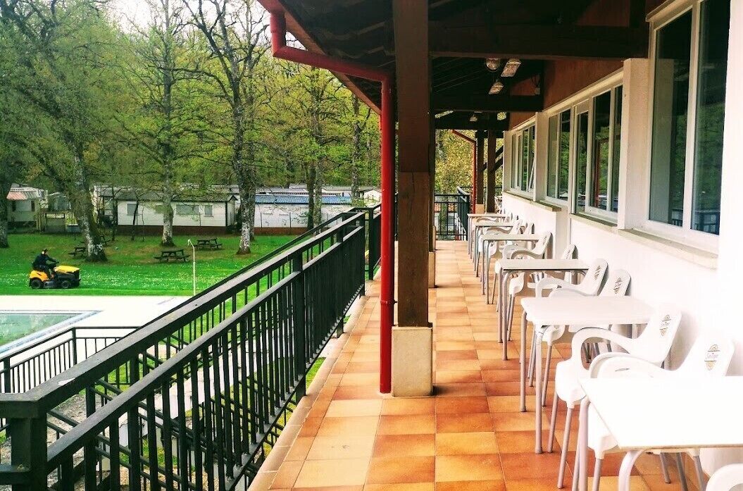 A covered outdoor area with a row of white tables and chairs, a black railing, and a view of a green park with trees and a yellow vehicle.