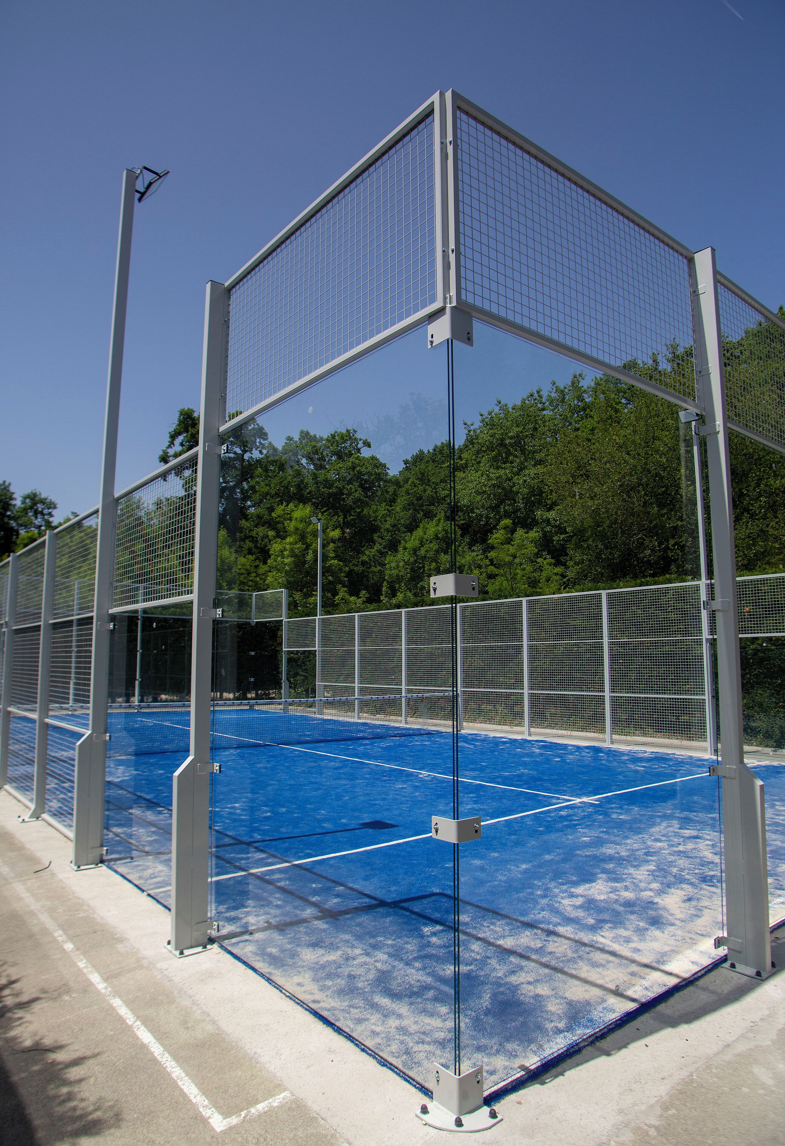 Instalación deportiva con cancha azul, cercado de metal y árboles al fondo.