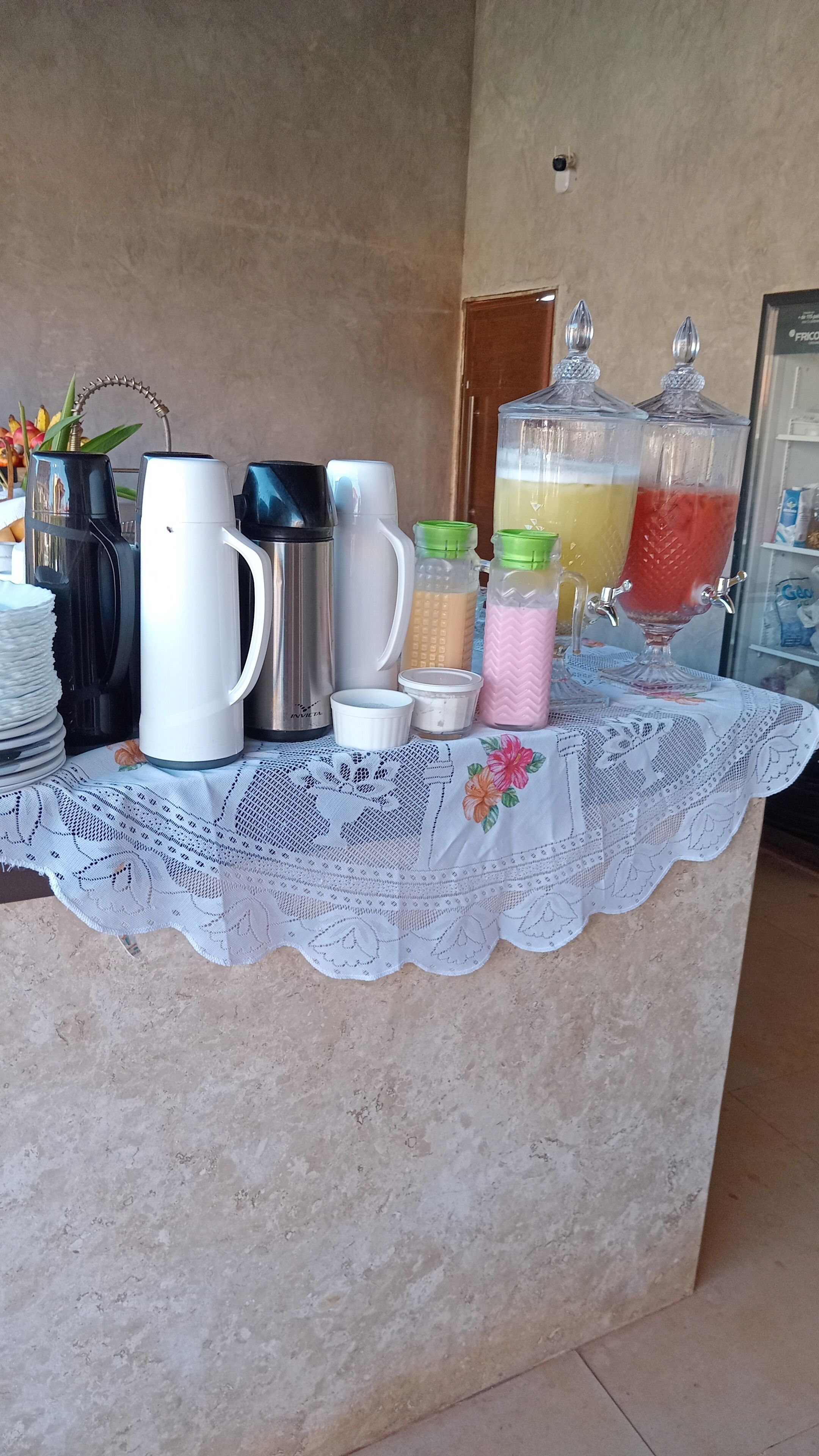 Montaje de un bufé de desayuno con varias máquinas de café, dispensadores de leche y un dispensador de ponche de frutas sobre una mesa cubierta con un mantel de encaje.