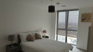 A bedroom with a large window, a bed with white bedding, a bedside table with a lamp, and a wall-mounted light fixture.