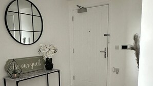 A hallway with a white door, a round mirror, a console table with a sign, and a vase of white flowers.