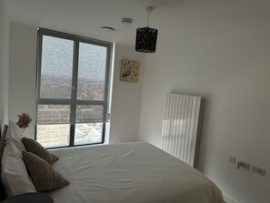 A bedroom with a large window, a bed with pillows, a hanging light fixture, and a radiator. - Immaculate 2-bed Apartment in East London (London)