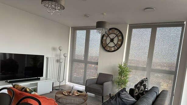 A modern living room with a large flat-screen TV, a grey sofa, a round coffee table, and a large window with a view.
