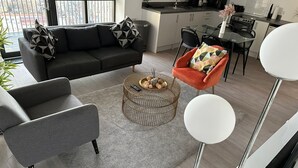 A modern living room with a black sofa, grey armchair, and a round coffee table with decorative items. A dining area with a glass table and chairs is visible in the background.