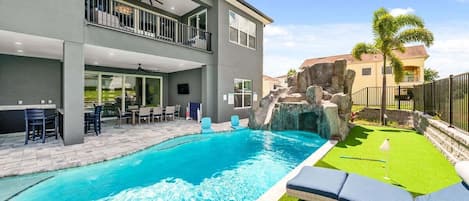 A modern two-story house with a swimming pool, outdoor seating, and a rock waterfall feature.