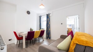 A modern living room with a sofa, dining table, and chairs. There is a window with curtains, a clock on the wall, and a door leading to another room. - Immaculate 3-bed House in Accrington (Accrington)