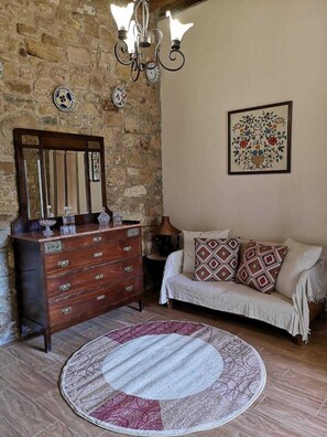 A room with a stone wall, a wooden dresser, a sofa with patterned cushions, a round rug, and a framed floral artwork. - M & M Stone House in Massari (Rhodes)