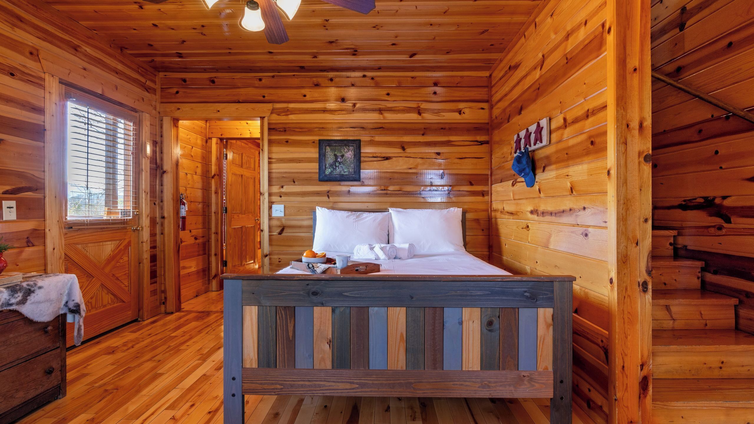 Quarto de cabana de madeira com uma cama, janela, porta e uma pequena mesa com toalha e frutas.