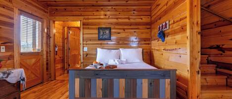 A wooden cabin bedroom with a bed, a window, a door, and a small table with a towel and fruit.