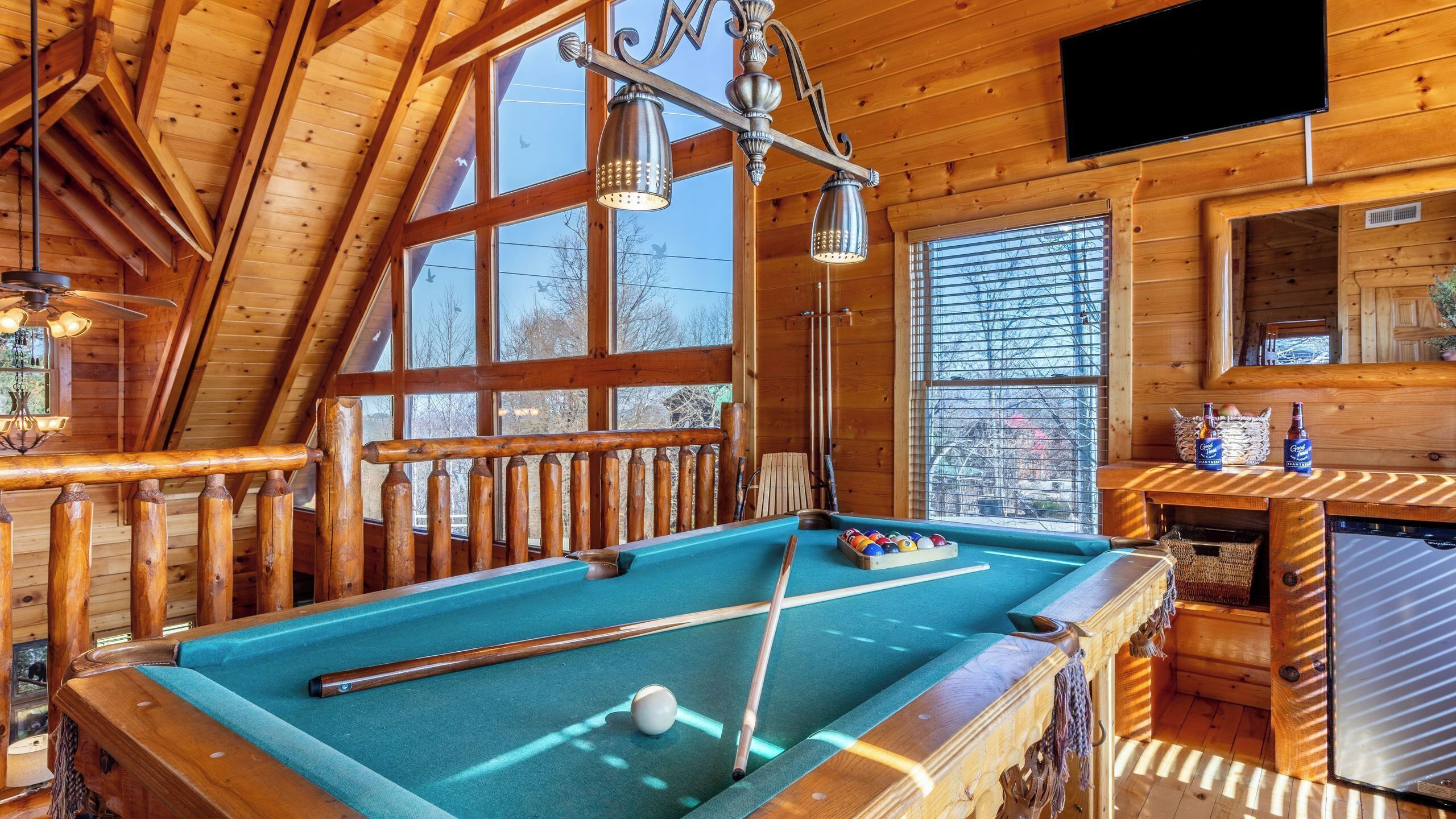 A pool table in a wooden room with a TV, wooden railings, and large windows.