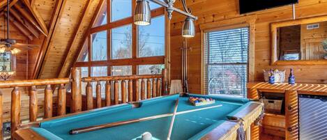 A pool table in a wooden room with a TV, wooden railings, and large windows.