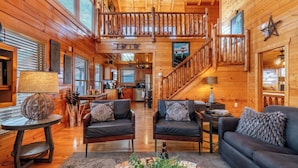 Un intérieur spacieux de chalet en bois, avec un coin salon central, une cuisine et un escalier menant à un niveau supérieur.