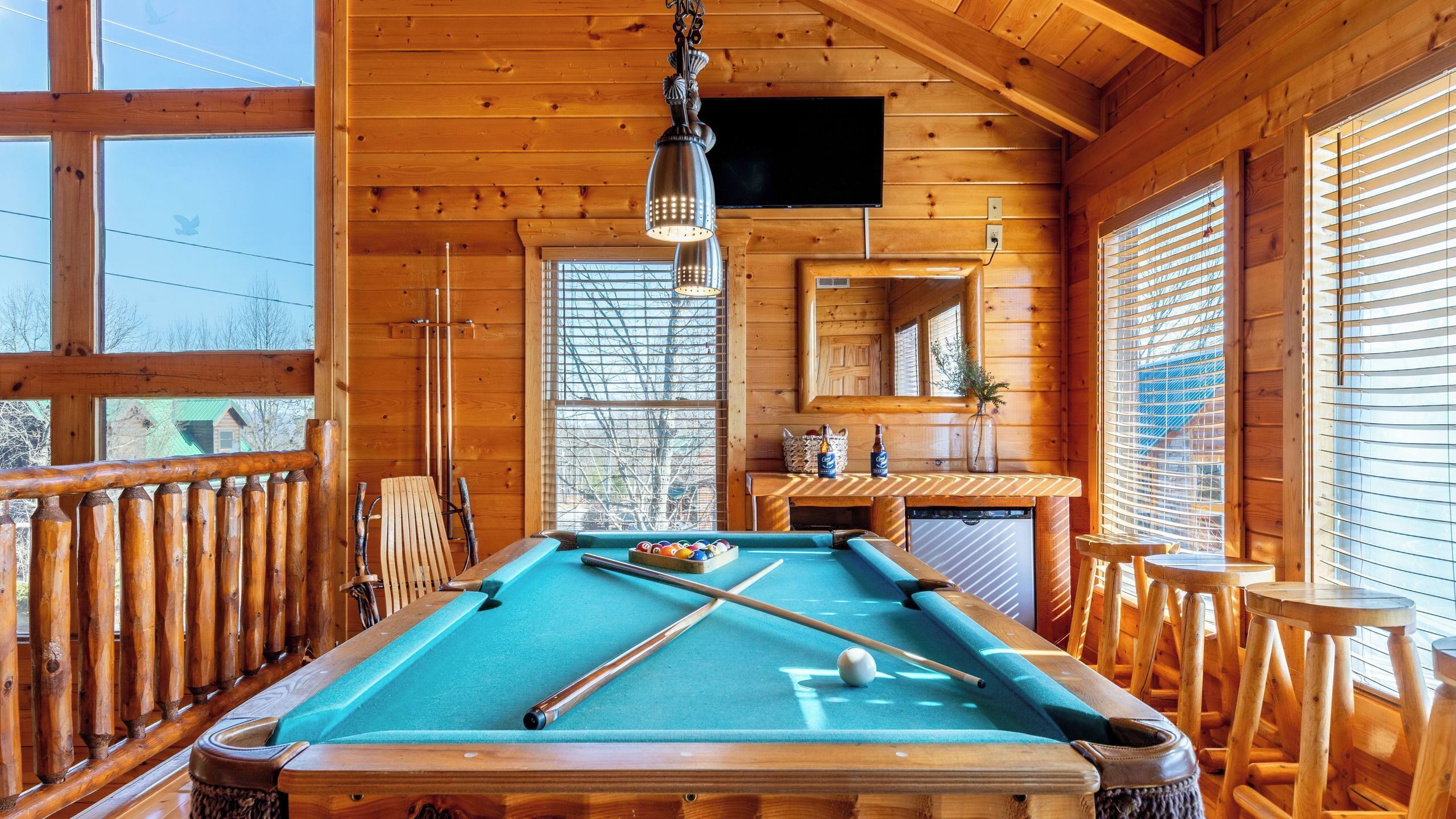 Una mesa de billar en una sala de madera con ventanas y televisor.