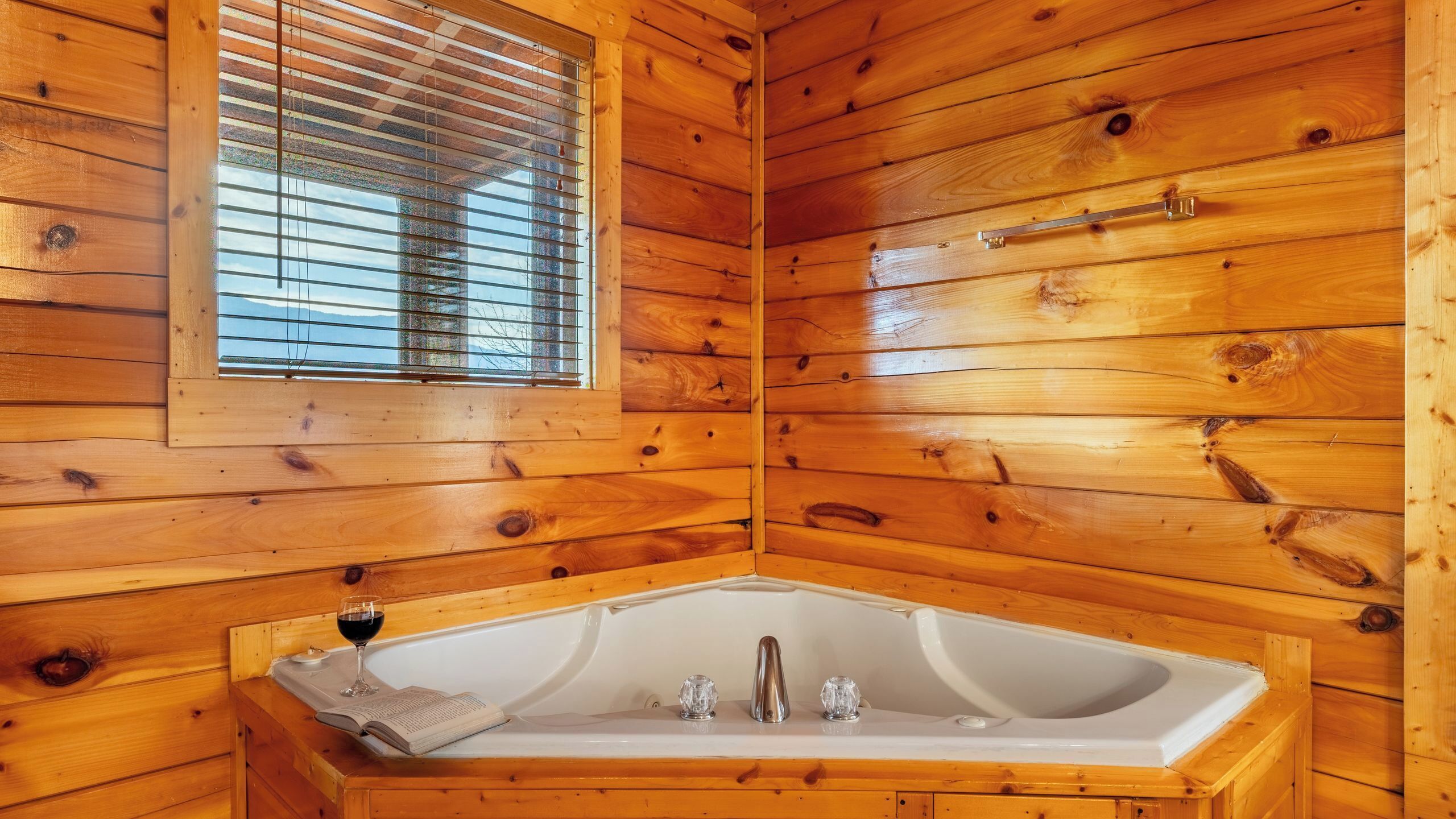 L’intérieur d’un chalet en bois, avec une baignoire d’angle, une fenêtre avec des stores et un verre de vin posé sur le sol.