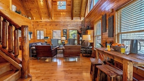 L’intérieur d’une cabane en rondins, avec un plancher en bois, une cheminée, un coin repas comprenant une table et des chaises, et un salon avec des canapés et une lampe.
