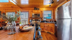 Une cuisine rustique aménagée dans une cabane en bois, avec un réfrigérateur en acier inoxydable, un four à micro-ondes et une cuisinière. On y trouve des placards en bois, une table en bois avec des assiettes à pain et des verres à vin, ainsi qu’une fenêtre équipée de stores.