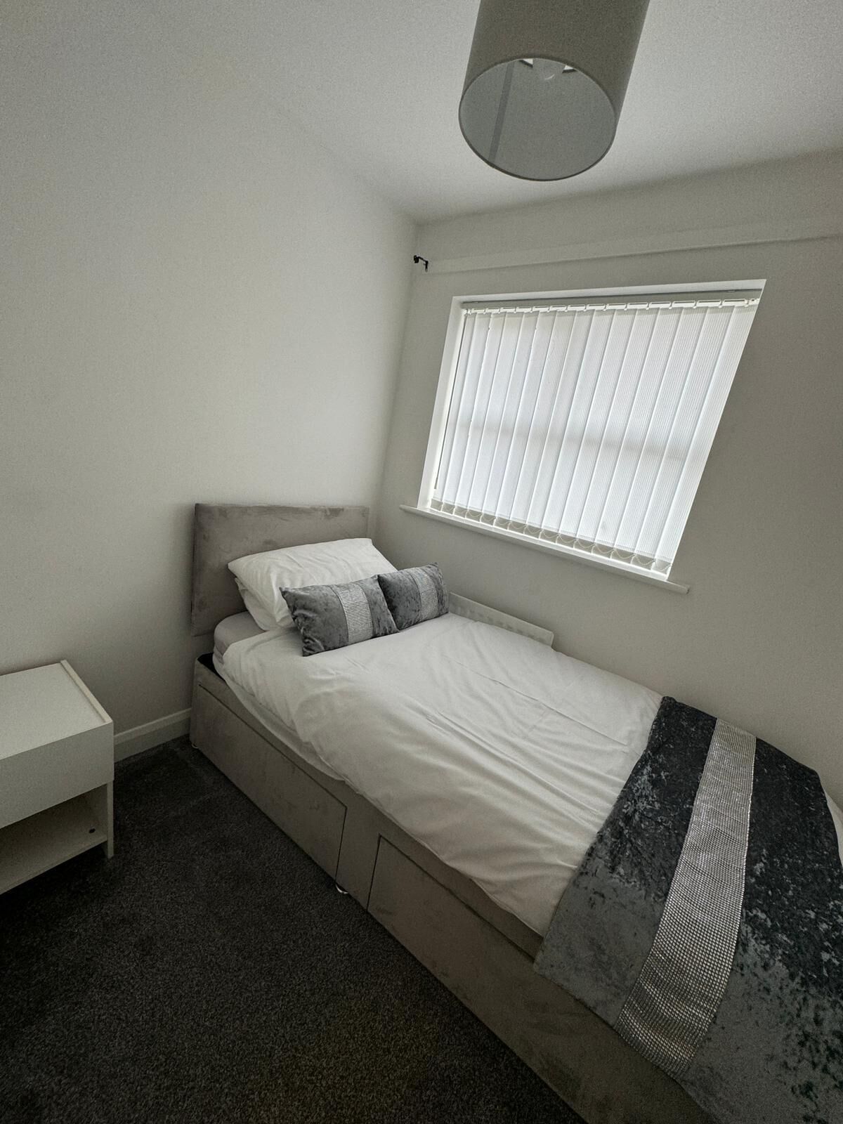 Una habitación pequeña con cama individual, cabecera gris, mesita de noche y ventana con persianas.