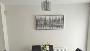 A dining area with a glass table, four black chairs, a vase with flowers, a textured pendant light, and a cityscape painting on the wall.