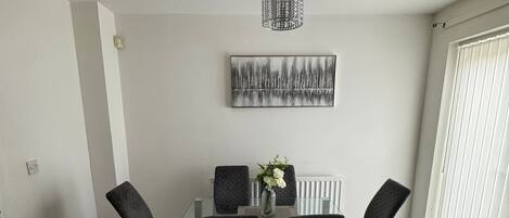 A dining area with a glass table, black chairs, a vase with flowers, a cityscape painting, and a textured light fixture.