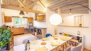Une cuisine moderne avec des poutres en bois, un coin repas avec une longue table dressée pour quatre personnes, et une bibliothèque contenant des livres et une plante.