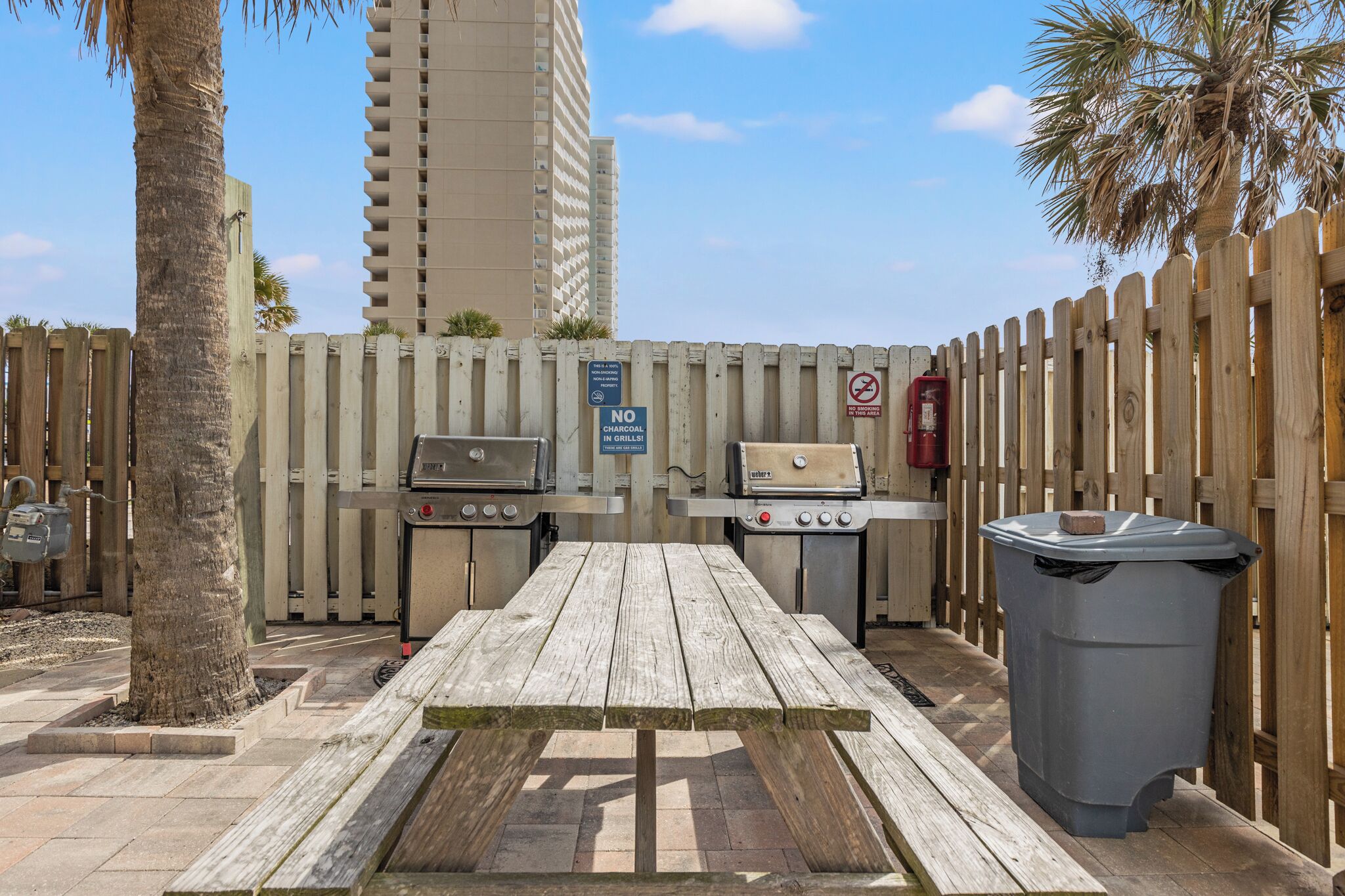An outdoor barbecue area with two grills, a picnic table, and a trash bin.