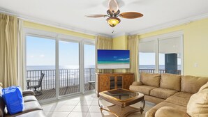 A living room with a large window overlooking the beach, a ceiling fan, and a flat-screen TV. - Grandview Oasis! 3BR 2BA Sleeps 9! Gulf-front Tranquility Beckons! (Panama City Beach)