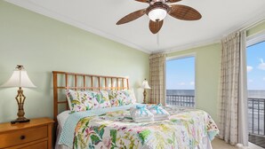 A bedroom with a bed, nightstands, a ceiling fan, and a view of the ocean. - Grandview Oasis! 3BR 2BA Sleeps 9! Gulf-front Tranquility Beckons! (Panama City Beach)