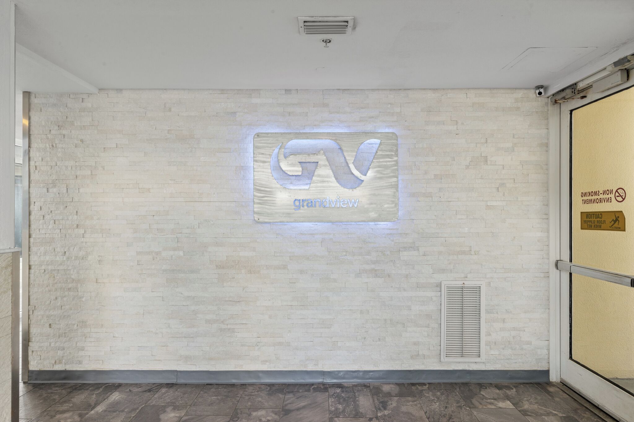 A reception area with a light-colored brick wall, a logo with the letters "GV" and the word "grandview" illuminated, a vent, and a door with a sign indicating no smoking and pet-friendly policy.