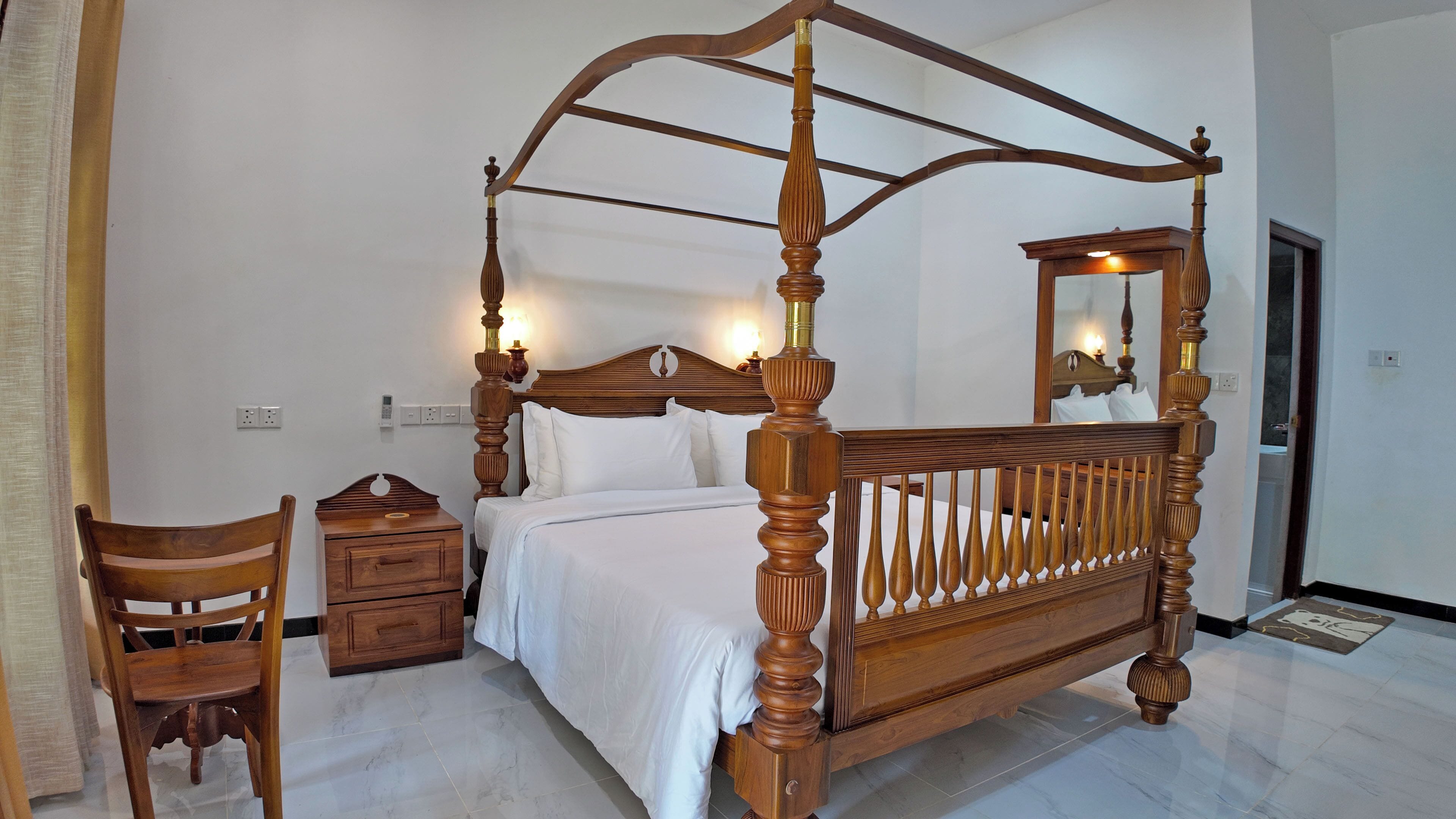 A four-poster bed with a wooden frame and a headboard with a curved design. There is a wooden nightstand with drawers and a chair next to the bed.