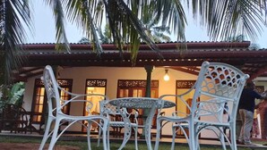 An outdoor seating area with white metal chairs and a table, surrounded by a garden and a building with a tiled roof. - Charika Villa (Bentota)