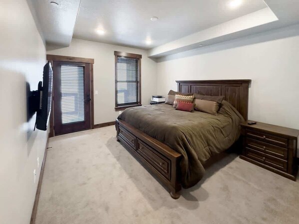 Quarto com uma cama grande, penteadeira, televisão na parede e uma porta que dá para outro cômodo.